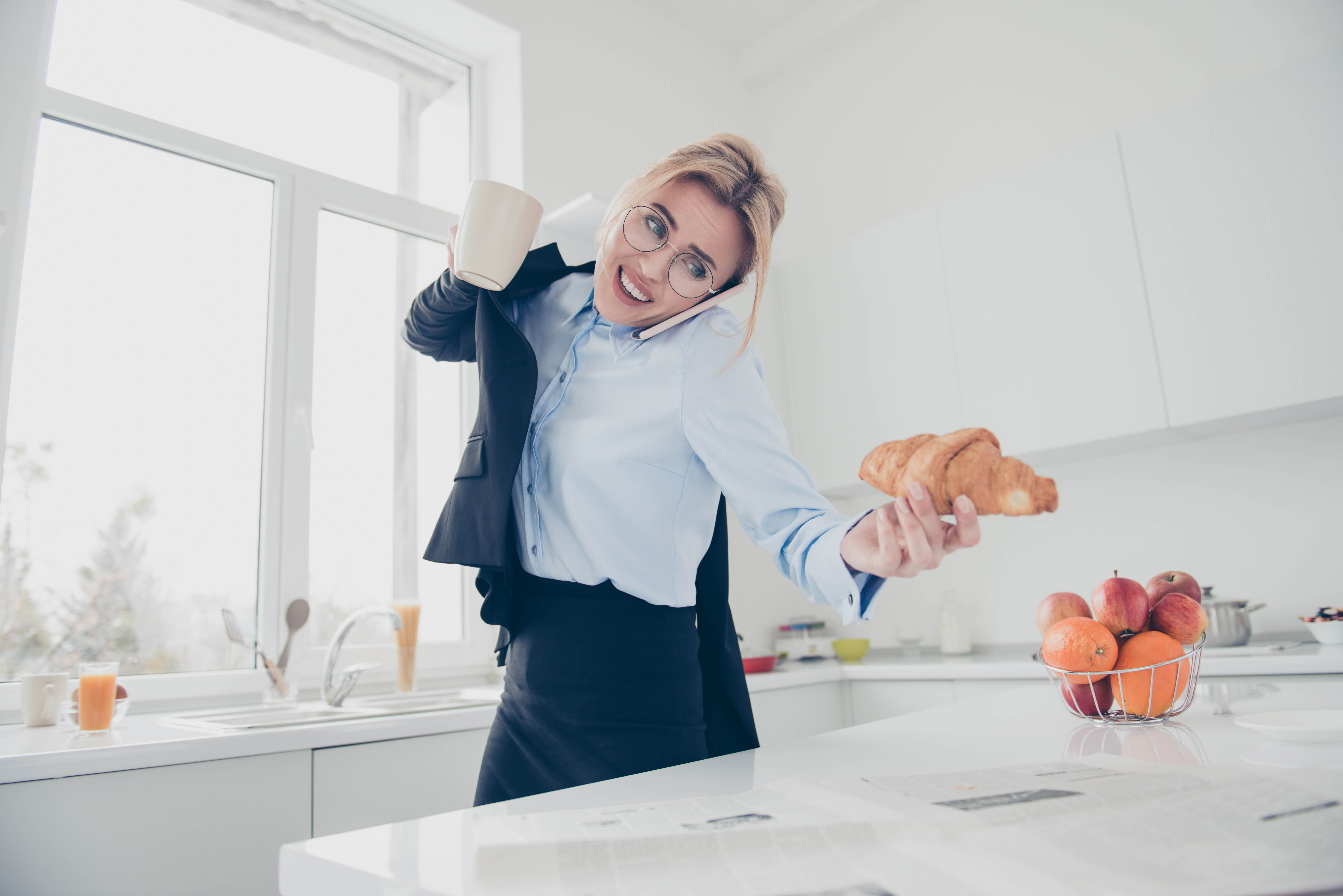 Blonde woman rushing in the morning, putting on her blazer while reaching for a croissant on the kitchen counter. She's holding a coffee cup in her top hand, that arm has her blazer already on.